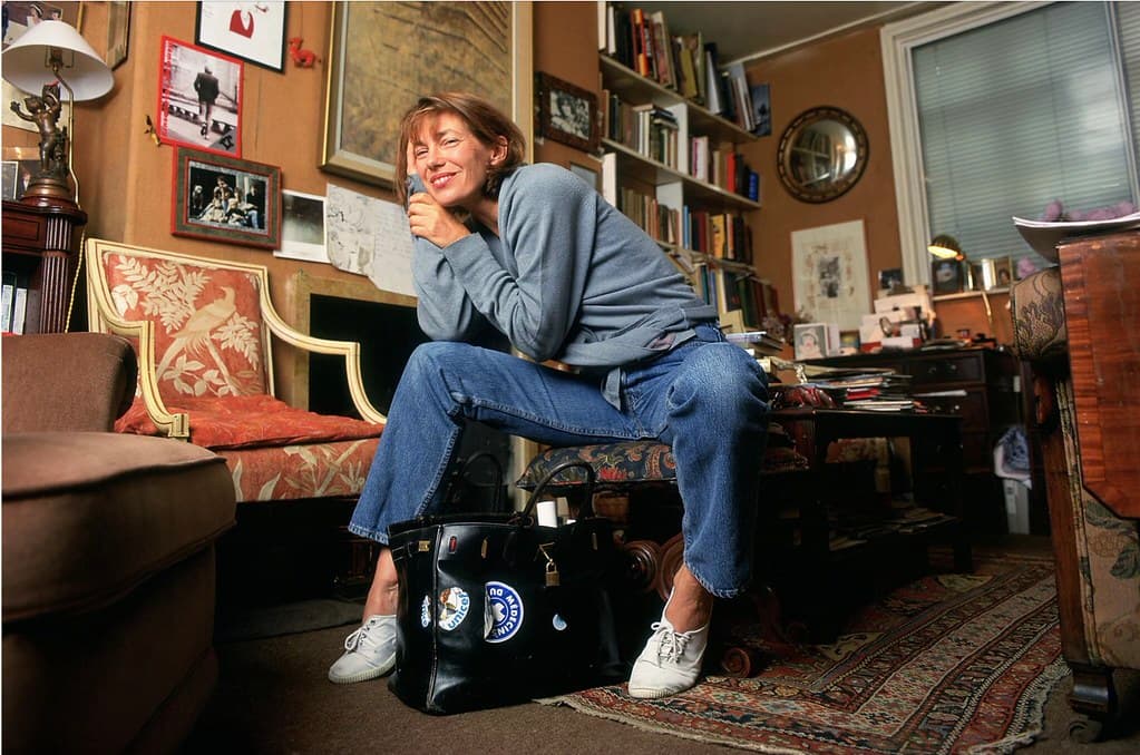 Jane Birkin sitting at home with her original black Birkin prototype handbag, showing natural wear and personal stickers.